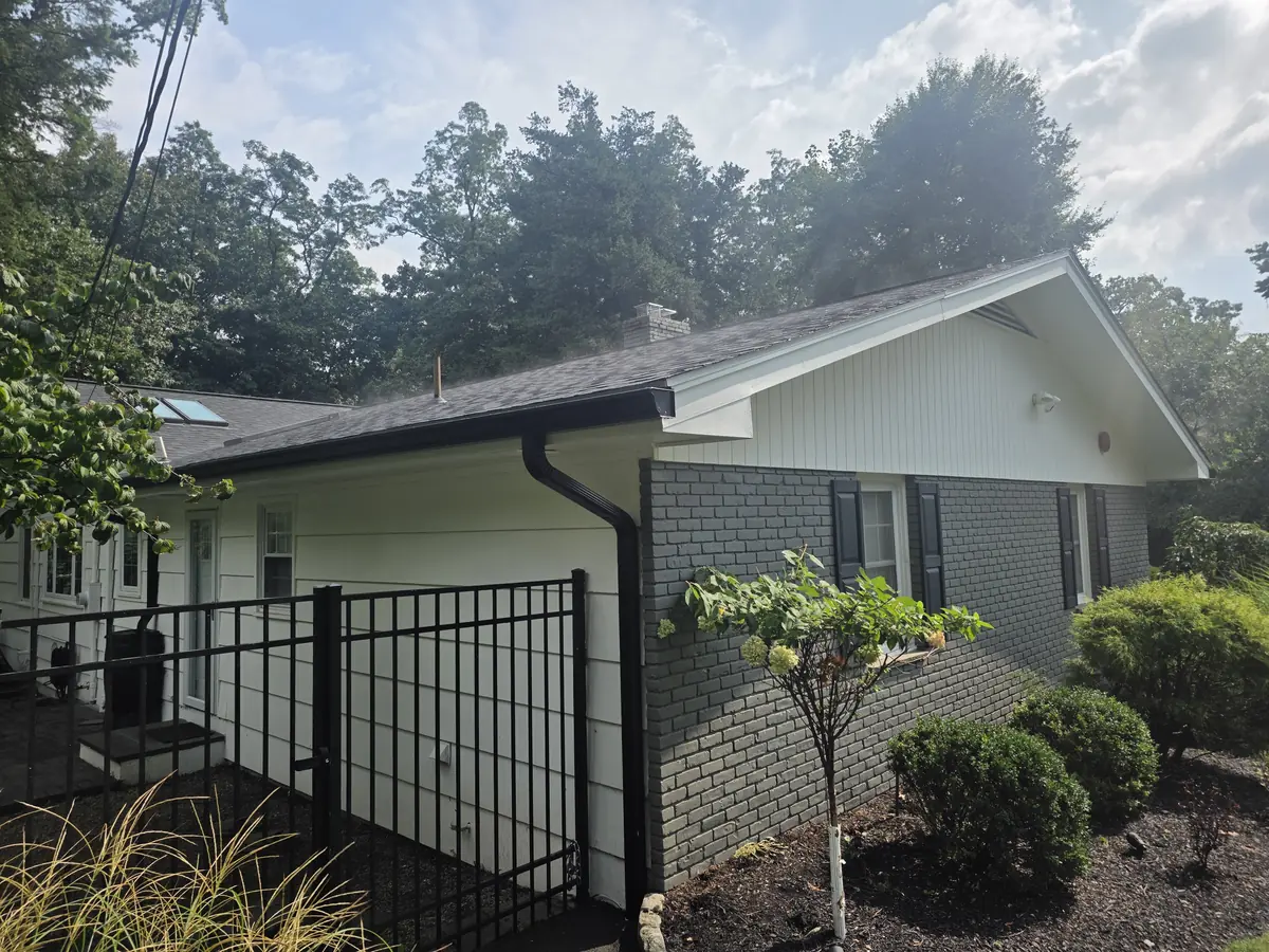 Side of a home under mature trees with a new black seamless gutter and downspout