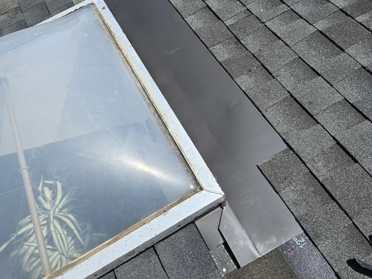 New dark metal flashing along the side of an older dome skylight, with the shingles finished along its outer edge