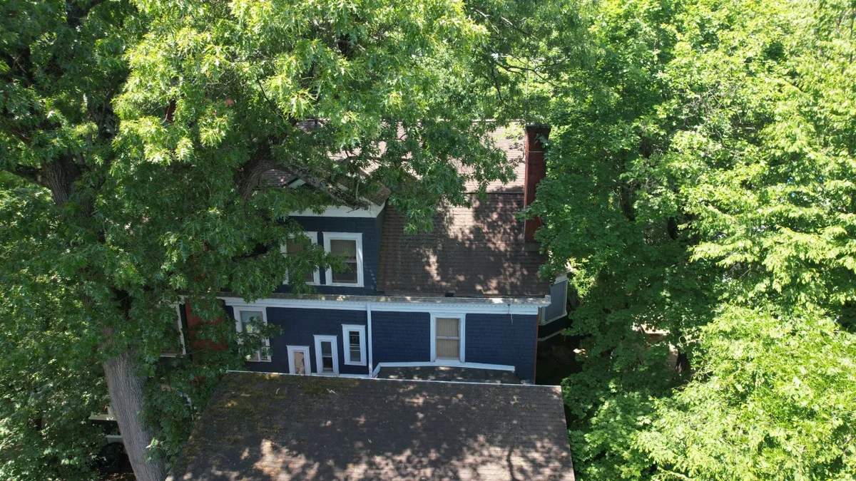 Aerial view of a dark blue older home with a brown shingle roof surrounded by mature trees