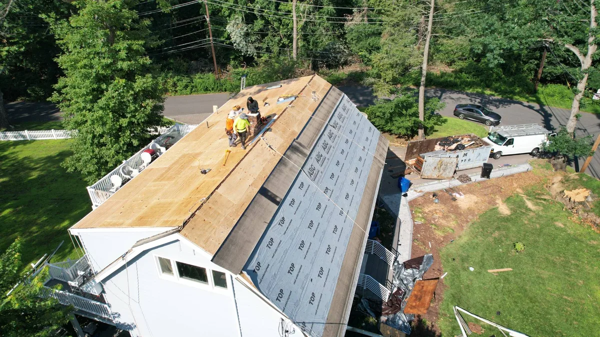 Aerial view of a house in the woods with new plywood roof decking and synthetic underlayment going down