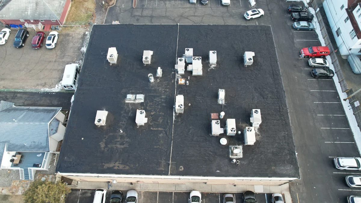 Drone photo straight down onto a large flat commercial roof in Little Falls, NJ: worn silver coating over gravel built-up roofing, dark patches, and about twenty rooftop heating and cooling units, with snow at the edges of the parking lot