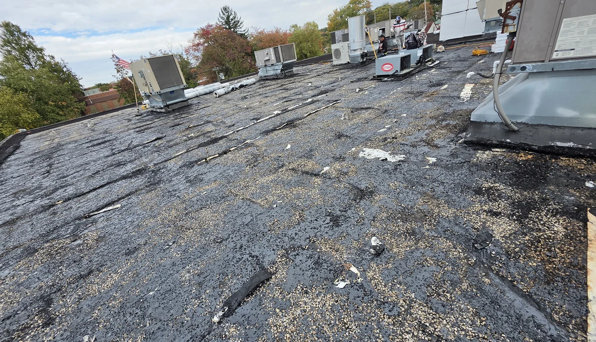 Close-up of an old commercial flat roof in Little Falls, NJ: gravel built-up roofing with the silver coating worn through, cracked and patched, rooftop units behind, and materials for the new roof staged at the far end