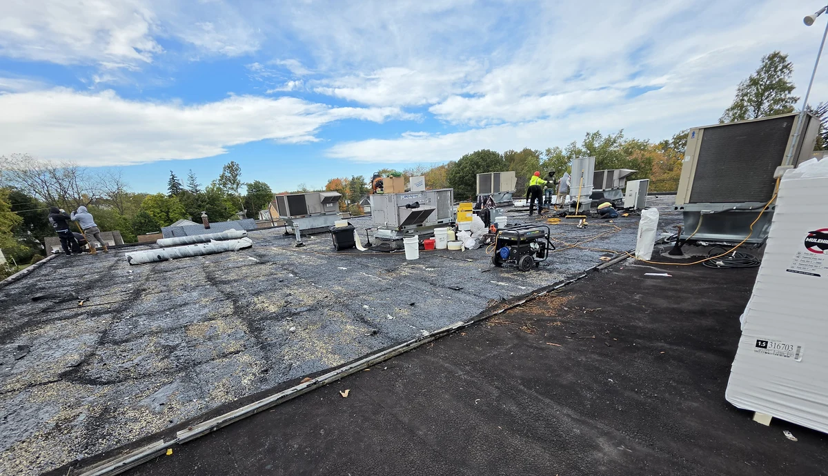 Commercial roof tear-off in progress in Little Falls, NJ: the old gravel built-up roofing on the left, the near section scraped down to the cracked black asphalt layers below, bundles of polyiso insulation and a generator, with the crew working beyond the rooftop units