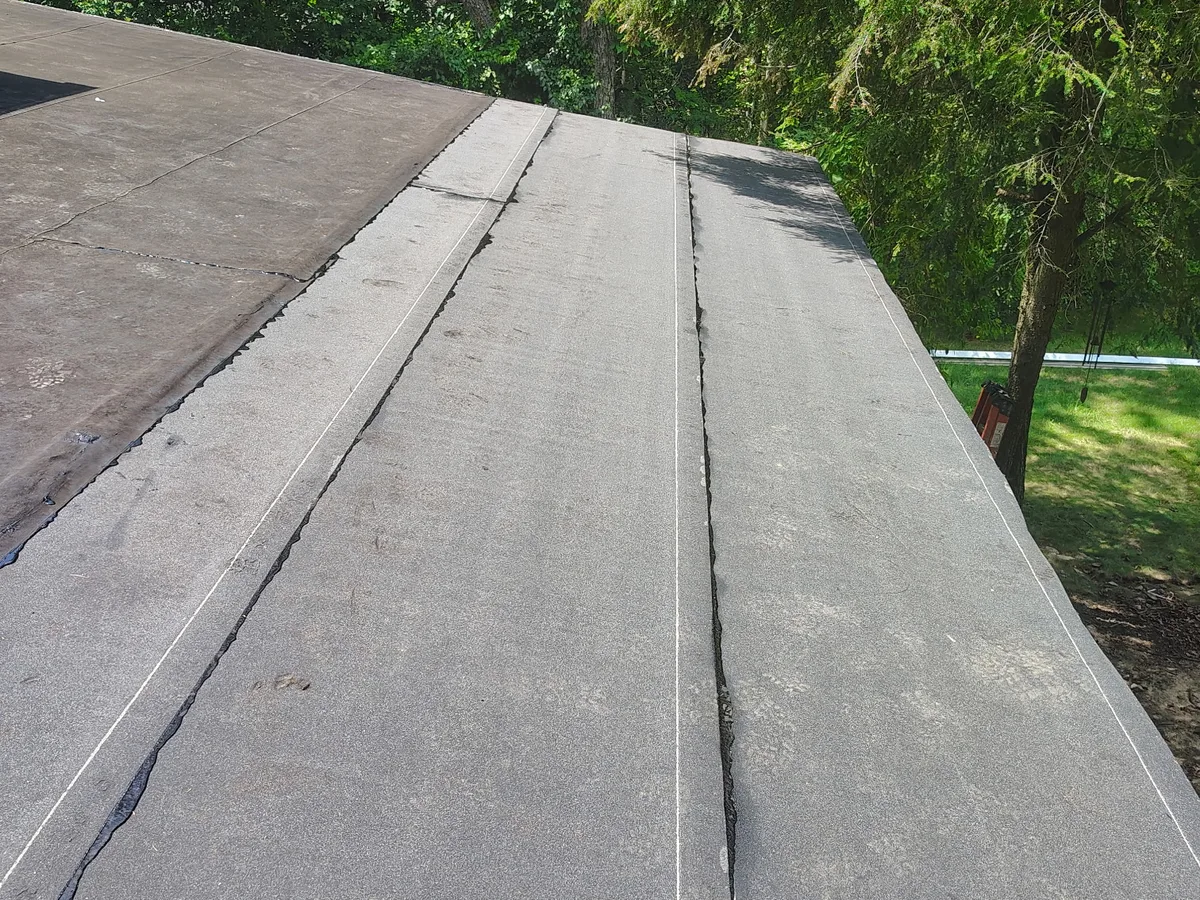 Granulated modified bitumen roof with every lap freshly re-sealed in black flashing cement, an older dark section on the left and trees beyond the roof edge