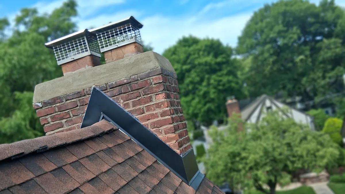 Brick chimney on a brown shingle hip roof with new black metal counter-flashing and a cricket behind it