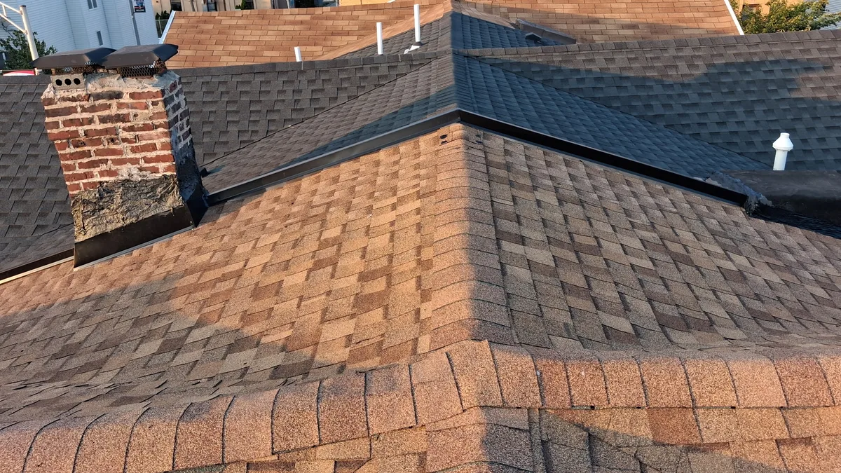 Low parapet wall between two shingle roof sections of one building, reframed and wrapped in new metal cap flashing, with a re-flashed brick chimney beside it