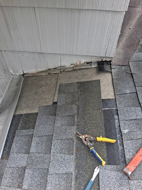 Old step flashing exposed where a gray shingle roof meets a shingle-sided wall, with the bottom edge of the siding damaged