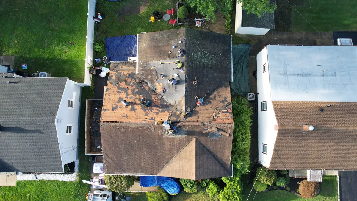Aerial view of a roofing crew stripping old shingles from a home, with the deck exposed and debris tarps below
