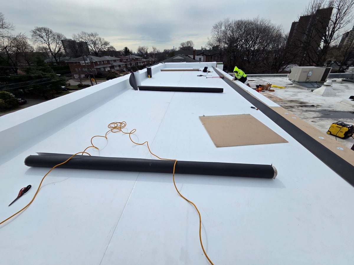 White TPO membrane sheets being rolled out and welded on a commercial flat roof, with a crew member working near the parapet