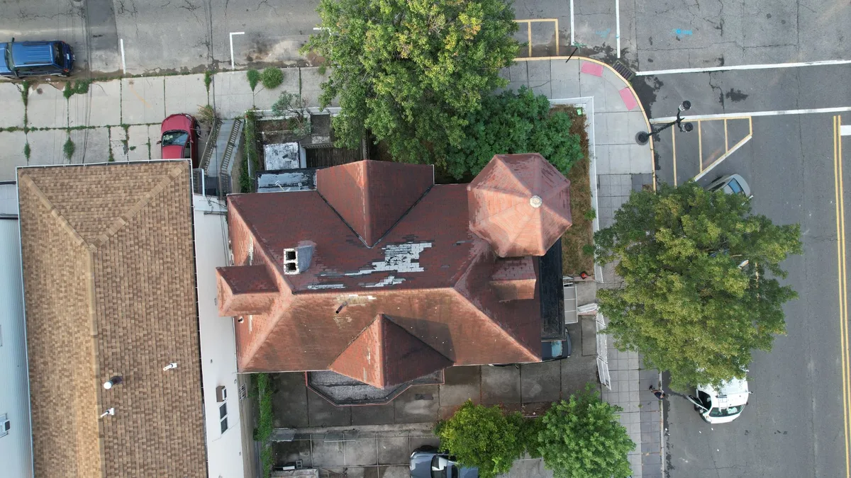 Aerial view of a Victorian home with a turret and a worn red tile-style roof with missing pieces